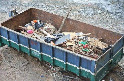 A large, rectangular metal skip with a rusted iron frame and blue-sided panels, positioned on a gravel surface in an outdoor setting. The skip contains a mixture of assorted construction and domestic waste including wooden planks, broken pieces of drywall or plasterboard, plastic bags, and various small discarded items. The materials appear weathered and are stacked haphazardly, with some pieces leaning against the sides of the skip. Surrounding the skip is a rough, uneven ground surface composed of gravel and dirt, typical of a waste collection or construction site. In the background, there is a gravel pathway or yard area, with no other objects or structures visible. The scene reflects a typical waste removal process, highlighting the type of rubbish managed by commercial waste services in Radlett, and emphasizes the importance of proper rubbish collection and disposal for local businesses and construction sites.
