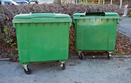 Two large green plastic wheeled rubbish bins are positioned side by side on a paved surface outdoors, likely in a commercial or residential area near Radlett. The bin on the left is slightly larger with a smooth, matte finish and a closed lid, while the smaller bin on the right has a similar finish and lid, with a few visible scratches and slight discolouration on the surface. Both bins are fitted with black plastic wheels at the base for mobility, and the lids are securely closed. In the background, there is a hedge with brownish foliage and some faint fencing, indicating an outdoor setting, possibly a driveway or service area. This scene relates to waste collection or rubbish management services, such as those provided by Commercial Waste Radlett, for waste removal on commercial or residential properties in the local postcode area. The overall lighting appears natural, suggesting daytime, with soft shadows cast on the ground. The image underscores the practical aspect of outdoor rubbish storage solutions within the Radlett community.