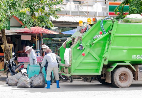 A street scene near a market or shopping area in Radlett, showing a bright green refuse collection truck positioned on the roadway with its rear compartment open, ready for waste collection. Two waste operatives wearing white protective suits, gloves, and blue footwear are actively handling large black and grey rubbish bags, which are placed on the pavement alongside the truck. The background features trees with green foliage, a small structure or stall with a red umbrella, and various market stalls or shops. The environment appears lively and typical of an urban area in Hertfordshire with some overhanging signage and partial building views, indicating a busy local space where rubbish removal services by Commercial Waste Radlett are actively part of maintaining cleanliness and waste management.