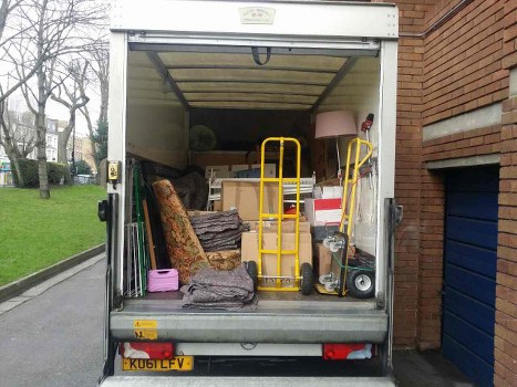 The image shows the open rear compartment of a white cargo van parked on a residential street near a brick building. Inside the van, various items of discarded household and furniture waste are visible, including a patterned armchair with fabric upholstery, a stack of flat-pack furniture or cardboard boxes, a pink plastic container, and a yellow hand truck leaning against other objects. Additional items include a small portable heater, a wooden lamp with a fabric shade, and miscellaneous office or household paraphernalia, all arranged in a somewhat cluttered manner. The background reveals a paved residential road with grass verges, mature leafless trees, and distant houses. The scene appears to be daytime with natural lighting, typical of a rubbish removal service in Radlett, indicative of the kind of waste that a business like Commercial Waste Radlett manages for clients requiring local rubbish clearance.