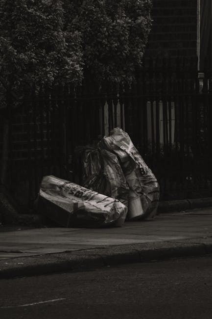 A collection of overflowing rubbish bins and scattered waste materials situated on a paved outdoor area, with various black, red, and grey bins filled with mixed paper, cardboard, plastic bags, and ot