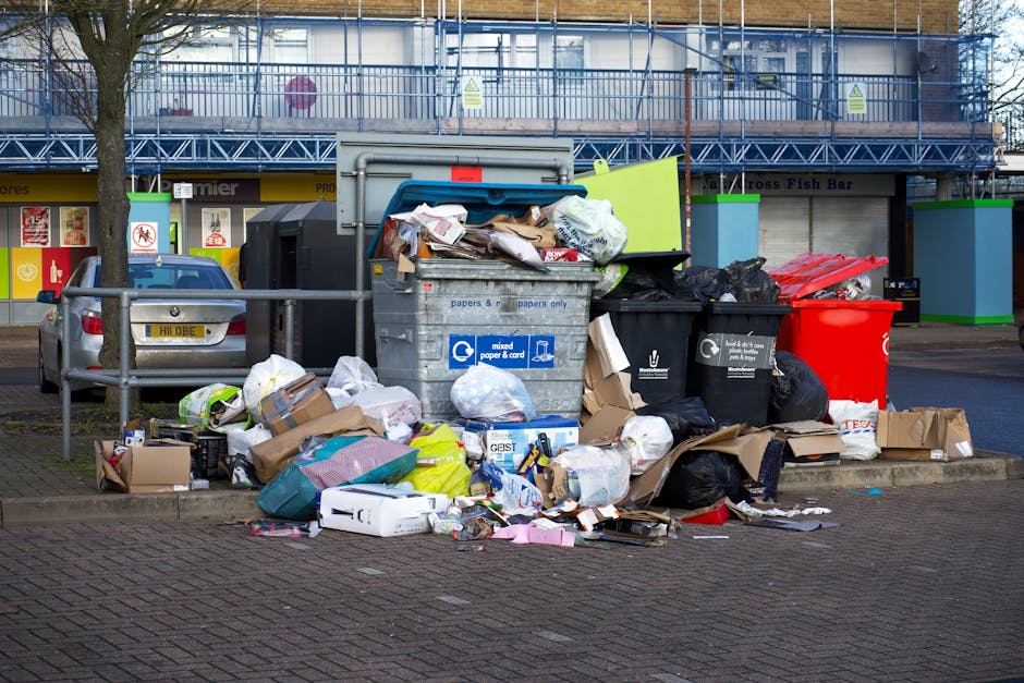 A middle-aged woman with short dark hair is seen leaning forward to dispose of rubbish into a silver, cylindrical urban litter bin with a rounded top and a front opening. She is wearing a black T-shir