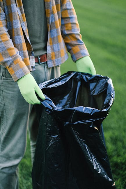 The image shows a person wearing a plaid shirt, grey trousers, and bright green gloves, standing outdoors on a grassy area. They are holding open a large black plastic rubbish bag, ready for waste col