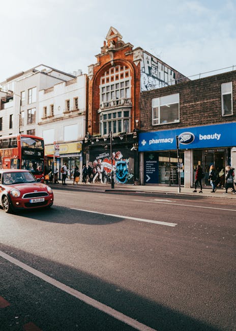 A busy urban street scene featuring a row of commercial buildings with a mix of architectural styles, including a prominent red-brick building with a large arched window on the upper floor and a decor